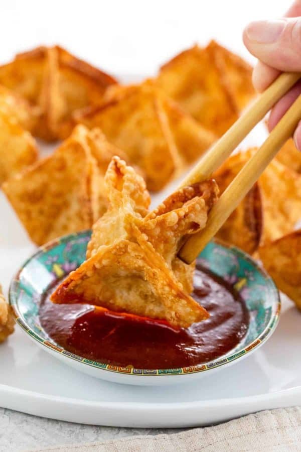 Person using chopsticks to dip crab rangoon in sweet and sour sauce.