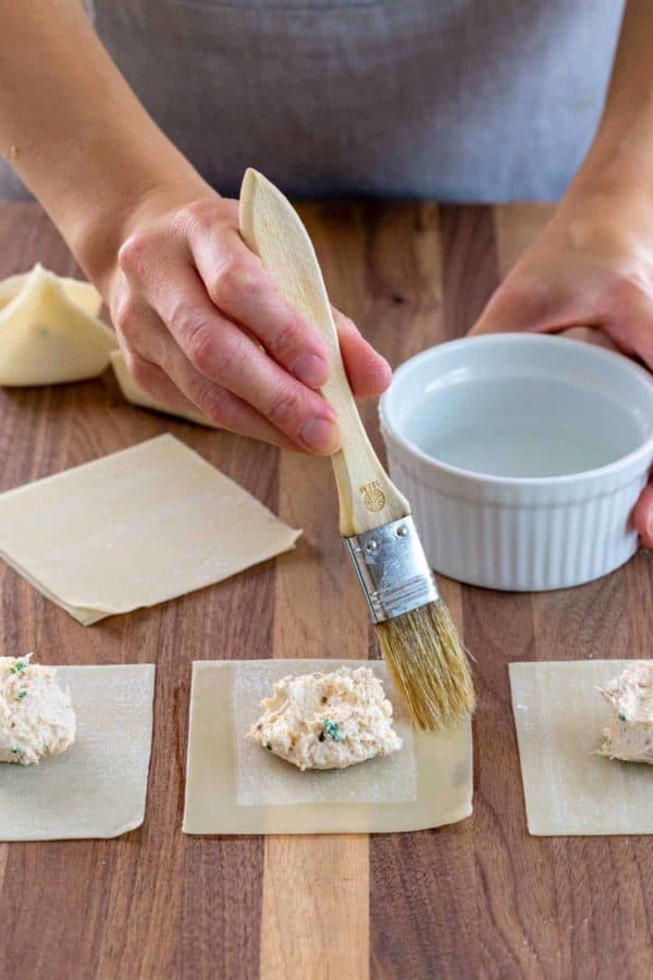 Person brushing water over the edges of a wonton wrapper with crab rangoon filling in the center.